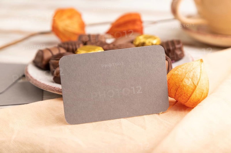 Gray business card with chocolate candies and physalis flowers on a white wooden background and orange textile. side view, close up, selective focus
