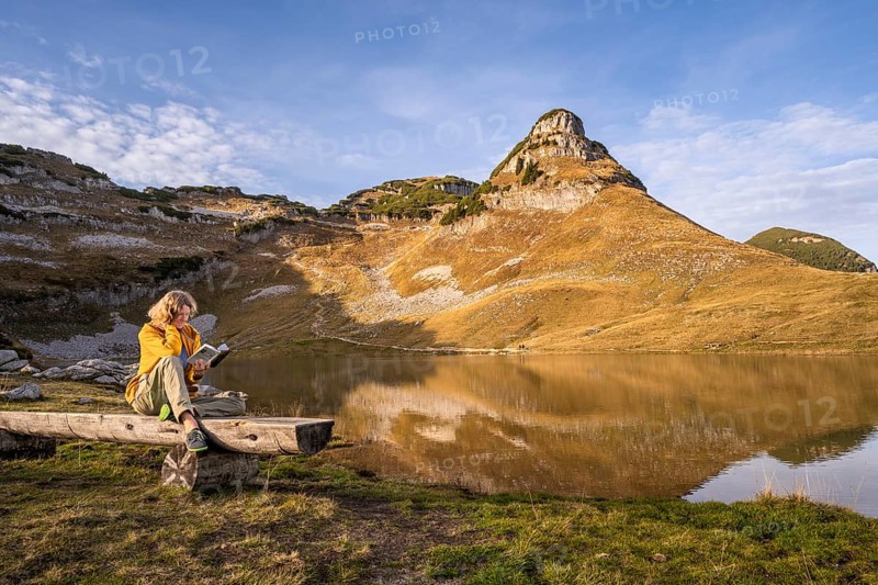 Lake Augstsee and the Atterkogel mountain on the Loser. A hiker sits on a bench and reads a book. Autumn, good weather, blue sky. Altaussee, Bad Aussee, Ausseer Land, Totes Gebirge, Styria, Upper Austria, Austria, Europe