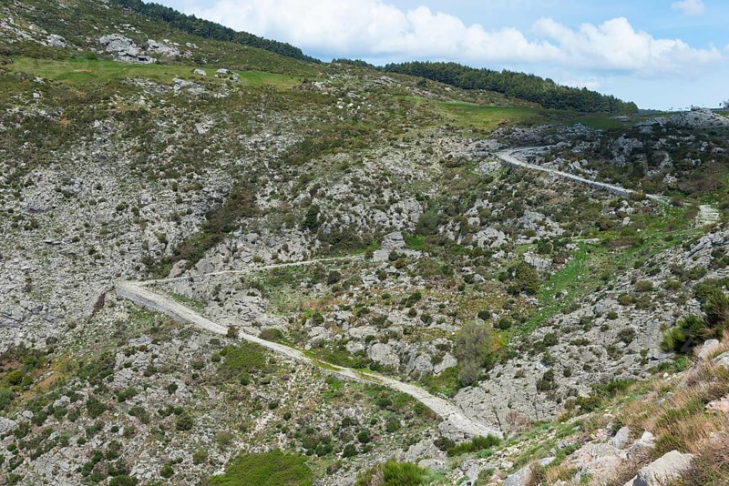 A winding serpentine road leads through a rocky mountain landscape with green grassy areas under a cloudy sky, old Roman road, at Puerto del Pico, Sierra de Gredos, Ávila, Avila, Castilla y León, Leon, Spain, Europe