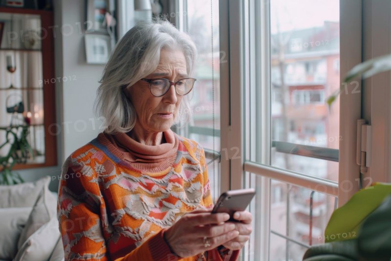 An elderly woman stands at the window in her living room and looks worried, frightened, unsettled at her smartphone, symbolic image for fraud against senior citizens, grandchild trick, shock calls, disclosure of personal data such as password, bank details, home address, media competence, AI generated, AI generated, AI generated