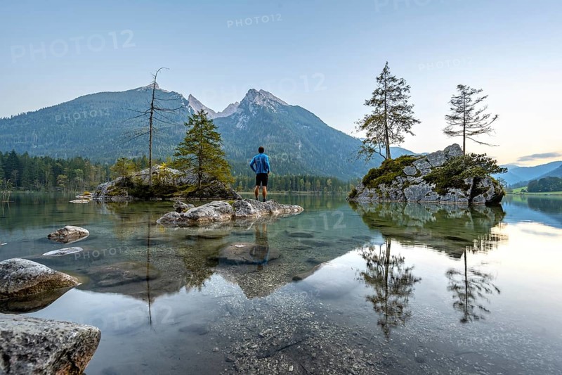 Young man standing on stones in the lake, Hochkalter reflected in Hintersee, at sunset, Berchtesgaden National Park, Ramsau, Upper Bavaria, Bavaria, Germany, Europe