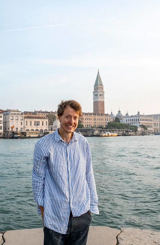 Young man in striped shirt and shorts on the banks of the Grand Canal, looking into the camera, Campanile in the background, Venice, Veneto, Italy, Europe