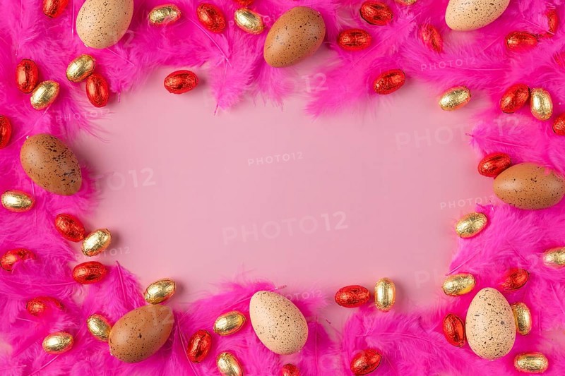 Frame made of eggs on pink feathers with red and gold chocolate eggs on a pink background
