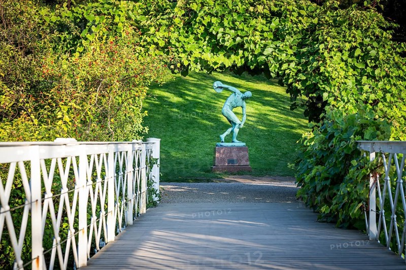 Wooden bridge leading to the sculpture of a discus launcher in a green, sunny park area, Botanical Garden or Botanisk Have, University of Copenhagen, Denmark, Europe