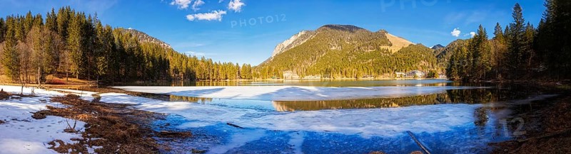Mountain lake partly covered with snow, surrounded by forests and mountains under a blue sky, Spitzingsee