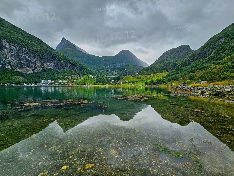 Mountainous landscape with a calm fjord reflecting the surrounding mountains and village, surrounded by green nature and under a cloudy sky, Geiranger, Geiranger Fjord, Stranda, Romsdal, Norway, Europe