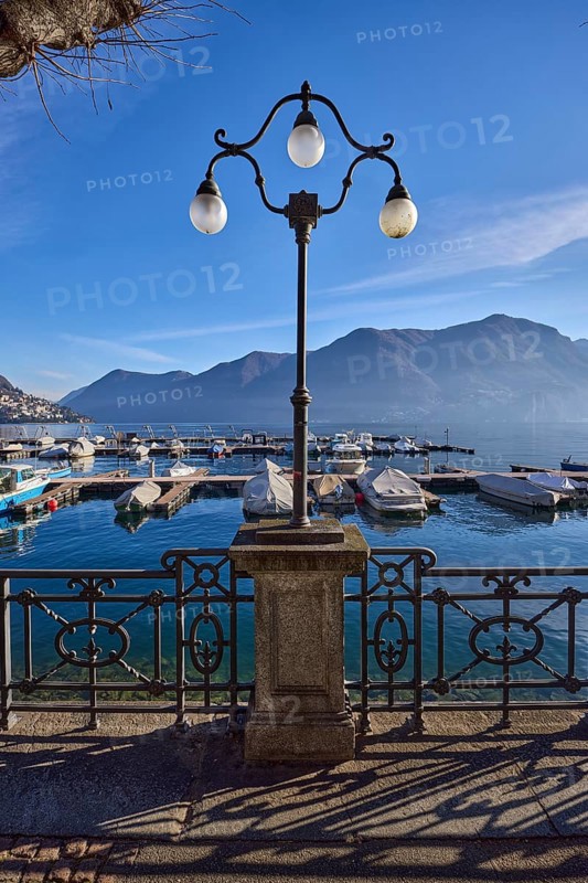 Lantern, forged railing, lakeside promenade, shadow, marina, tree, motorboats, haze, mountain, backlight, blue sky with veil clouds, Lake Lugano, Lugano, district Lugano, canton Ticino, Switzerland, Europe