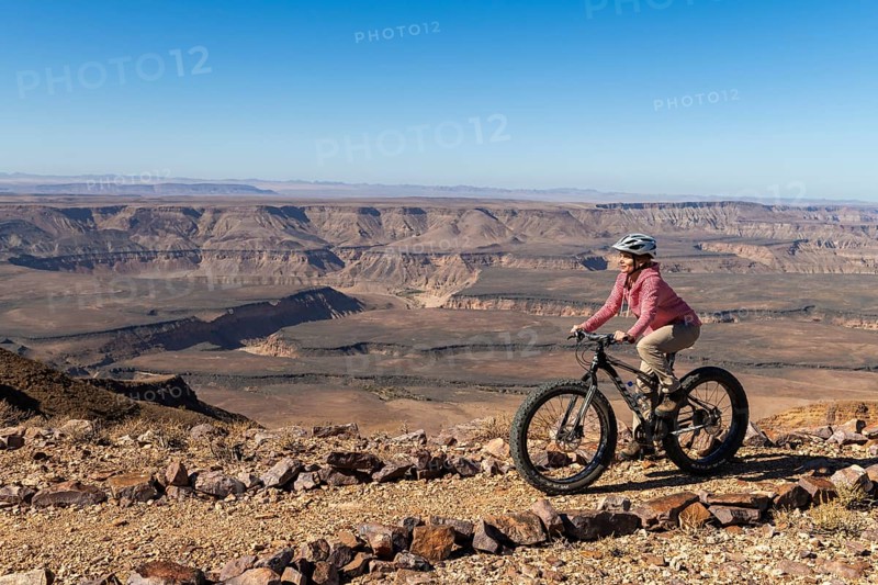 Woman riding a mountain bike, fatbike, on the edge of the abyss, behind the gorge landscape of the Fish River Canyon, Fish River Gorge, Namibia, Africa