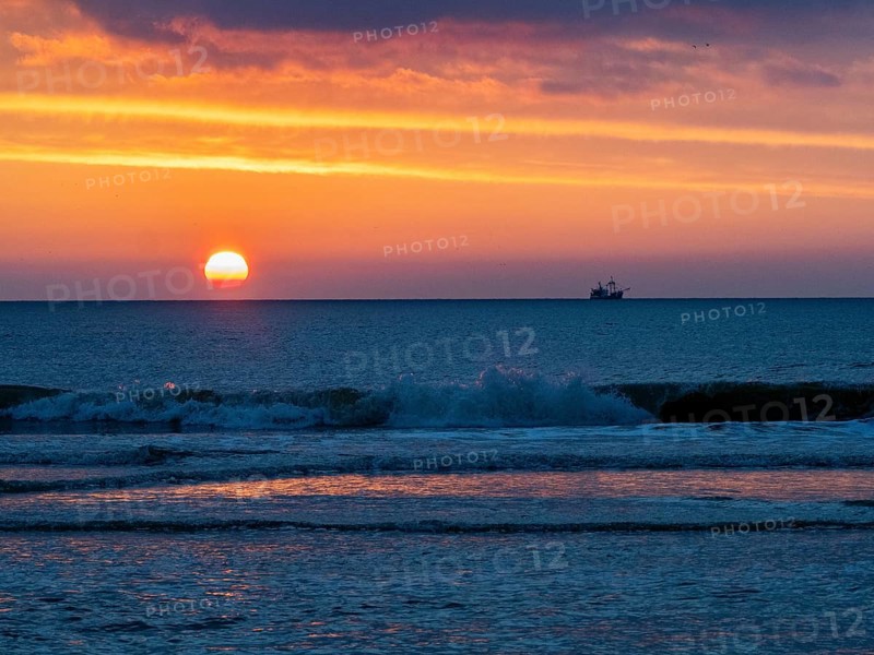 Silhouette of a fishing boat against a colorful sunset over the North Sea, and waves breaking on the beach in the foreground, on the West coast from the island of Texel, Holland