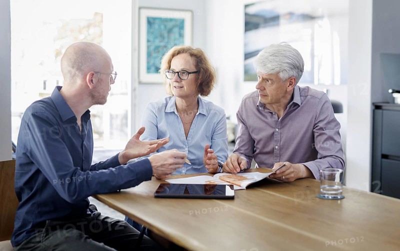 Symbolic photo on the subject of counselling. An older woman and an older man sit together at a table at home and receive counselling from a young man. Berlin, 13.08.2024