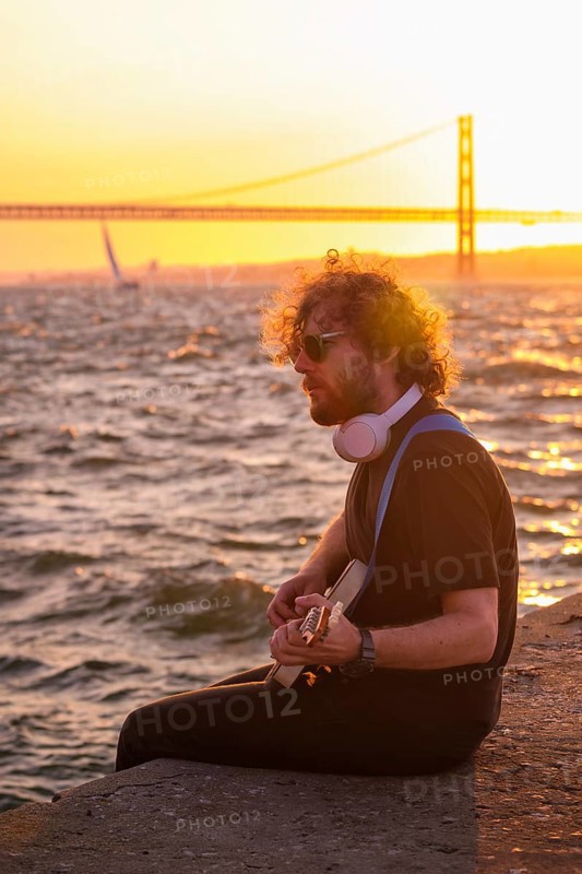 Hipster street musician in black playing electric guitar in the street on sunset on embankment with 25th of April bridge in background. Lisbon, Portugal, Europe