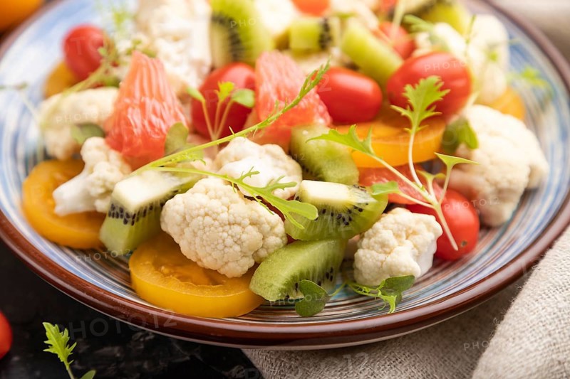 Vegetarian salad of cauliflower cabbage, kiwi, tomatoes, microgreen sprouts on black concrete background and linen textile. Side view, close up, selective focus