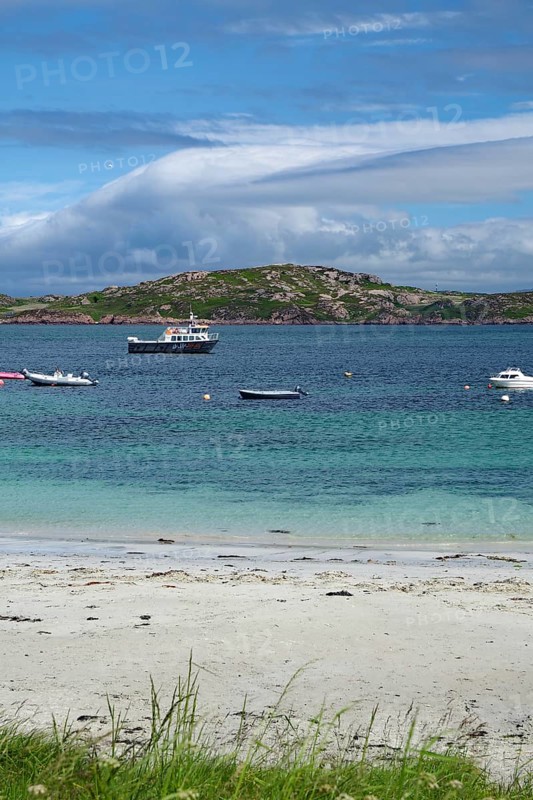 Boats in blue water in front of an island, with white clouds and a beach in the foreground, Holy Island, Iona, Inner Hebrides, Scotland, Great Britain
