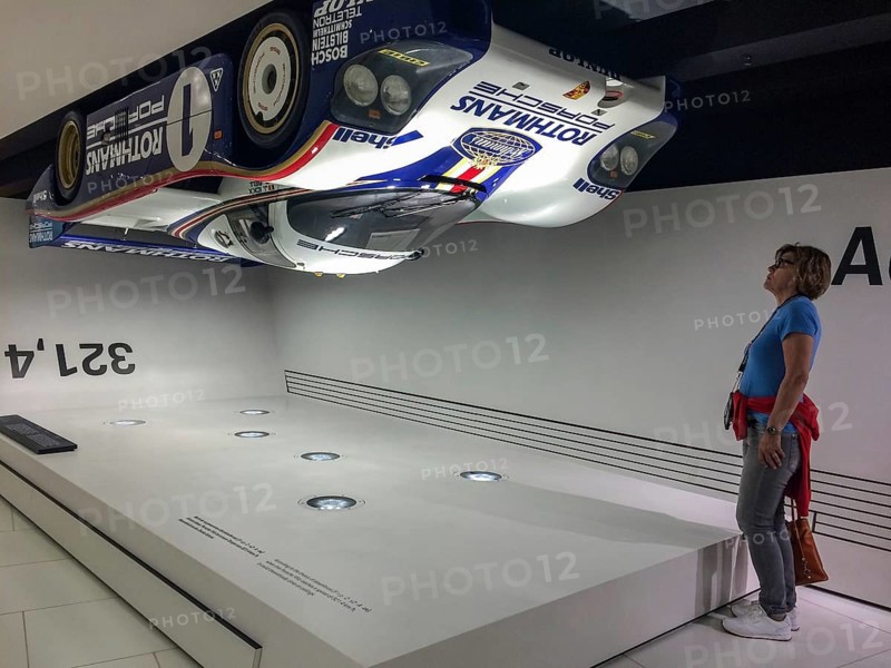 Visitor to the museum looks upwards at a Porsche 956 racing car hanging upside down under the ceiling of the museum as an example of high contact pressure at 321.4 kilometres per hour, Porsche Museum, Stuttgart, Baden-Württemberg, Germany, Europe
