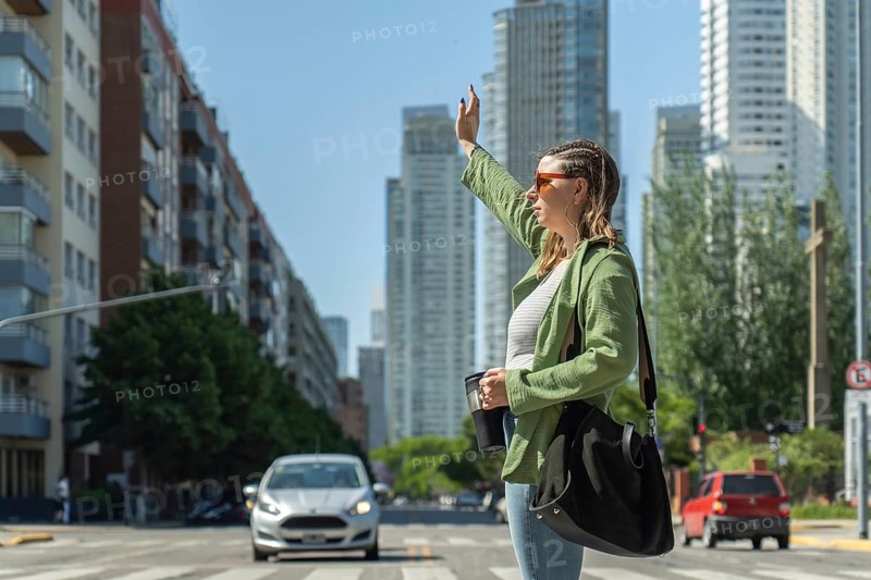 Young woman with sunglasses and a shoulder bag is hailing a ride on a city street, holding a reusable coffee cup while cars pass by and skyscrapers rise in the background