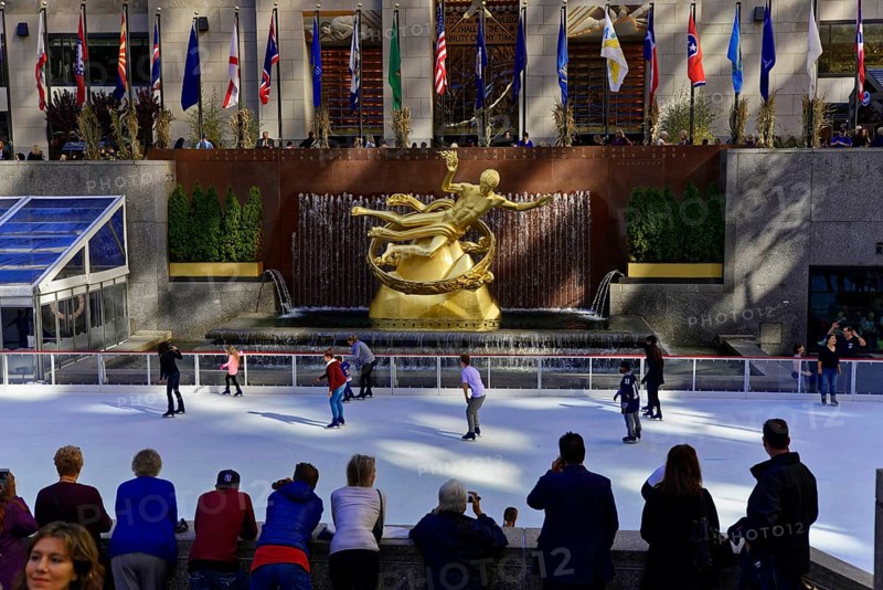 Rockefeller Center observation deck, ice skaters in front of the Prometheus sculpture with flags hanging above in a busy city, Manhattan, New York City, New York, USA, North America
