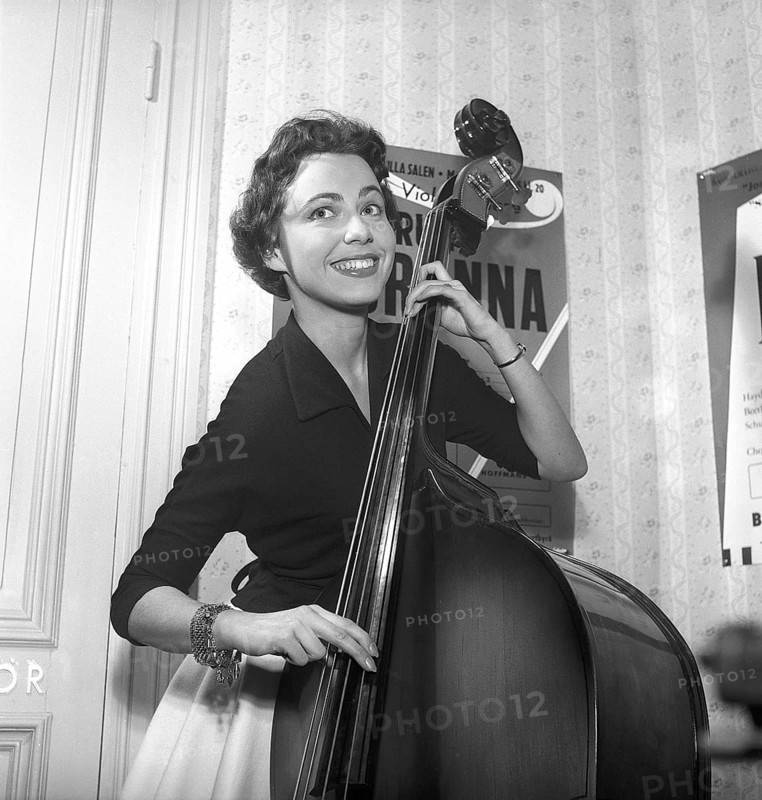 Girl in 1956. The photograph, taken in 1956, portrays a young woman with a bright smile as she plays a double bass. She stands confidently, her fingers deftly plucking the strings of the towering instrument, her joyful expression radiating enthusiasm. The image captures a moment of musical passion, framed by the mid-1950s aesthetic, with the double bass adding a bold, resonant presence to her cheerful demeanor. Kristoffersson ref BS3-5
