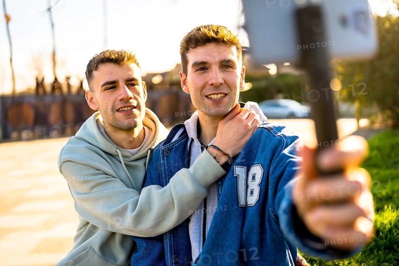 Two men in love embracing each other while capturing a joyful selfie with a smartphone mounted on a gimbal in a sunny urban park