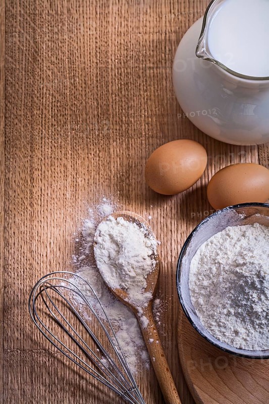 Aerial view copyspace composition pitcher with milk flour in bowl and spoon eggs corolla on wooden board food and drink concept