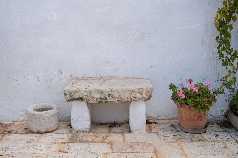 Stone bench and flower pot with geraniums on a rustic stone floor in front of a white wall, Apulia, Italy, Europe