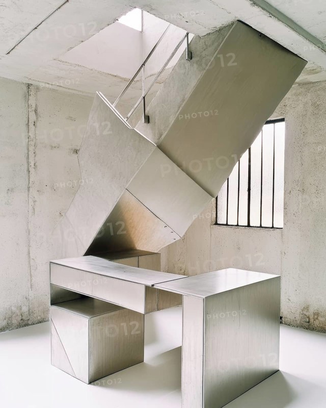 The image features a bright, minimalist interior with a modern desk constructed of metallic material. An overhead structure with visible supports complements the design. Natural light streams through the window, AI generated