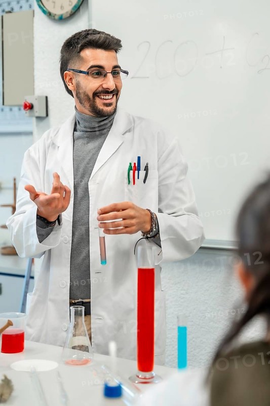 Smiling chemistry teacher showing colorful test tubes and beakers while explaining chemical experiment to students in a university laboratory