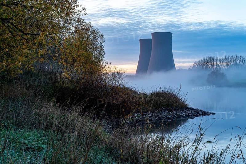 Cooling towers of the Grohne nuclear power plant in the fog in the morning, surrounded by autumnal riverbank of the Weser and trees creating a mysterious atmosphere, Grohnde, Lower Saxony, Germany, Europe