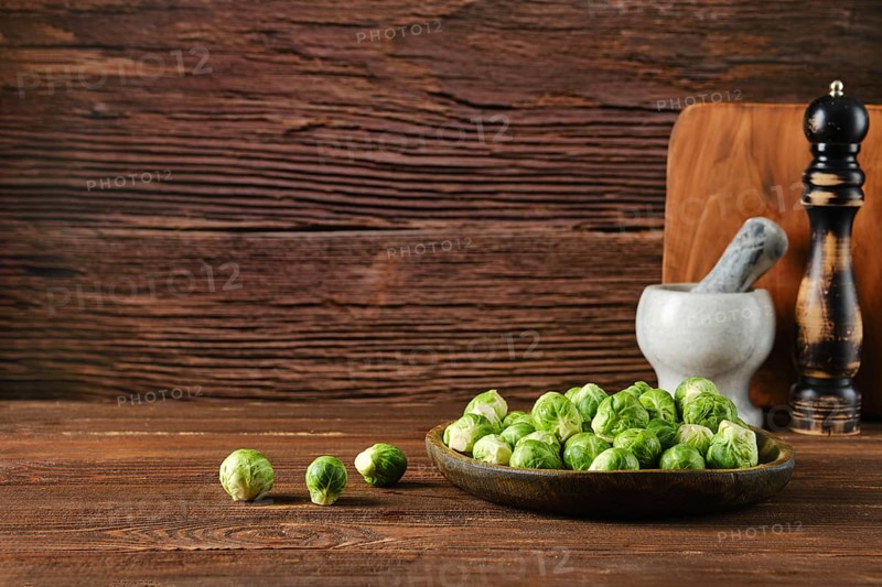 A wooden bowl filled with fresh Brussels sprouts sits on a rustic wooden table. Nearby, a mortar, pestle, and pepper mill add to the kitchen ambiance, emphasizing the ingredients\' freshness.