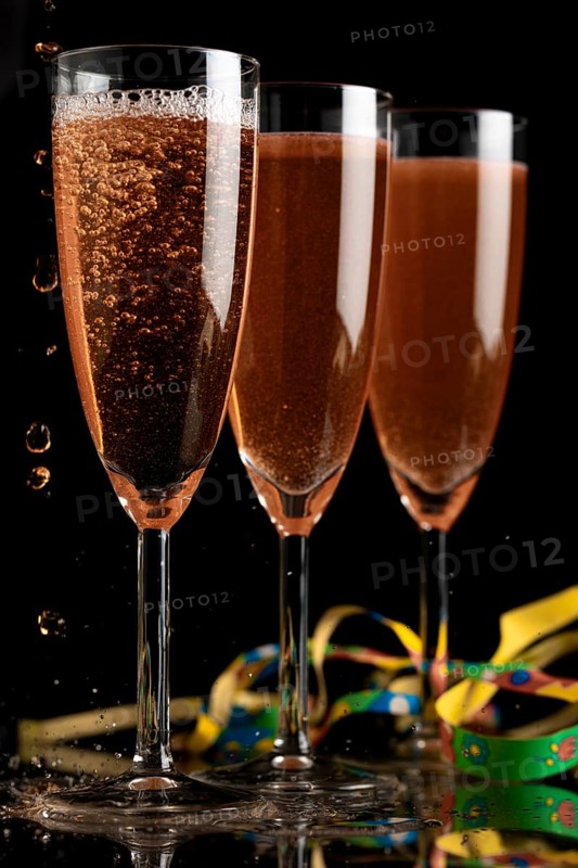 Three champagne glasses on a dark background, liquid bubbling, colourful ribbons next to them
