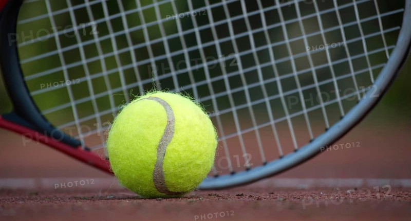Close up of a yellow tennis ball and a tennis racket lying on a tennis hard court surface is depicted