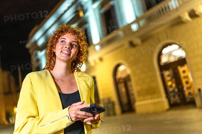 Happy woman in casual colorful clothes and curly hair looking at city views using phone at night