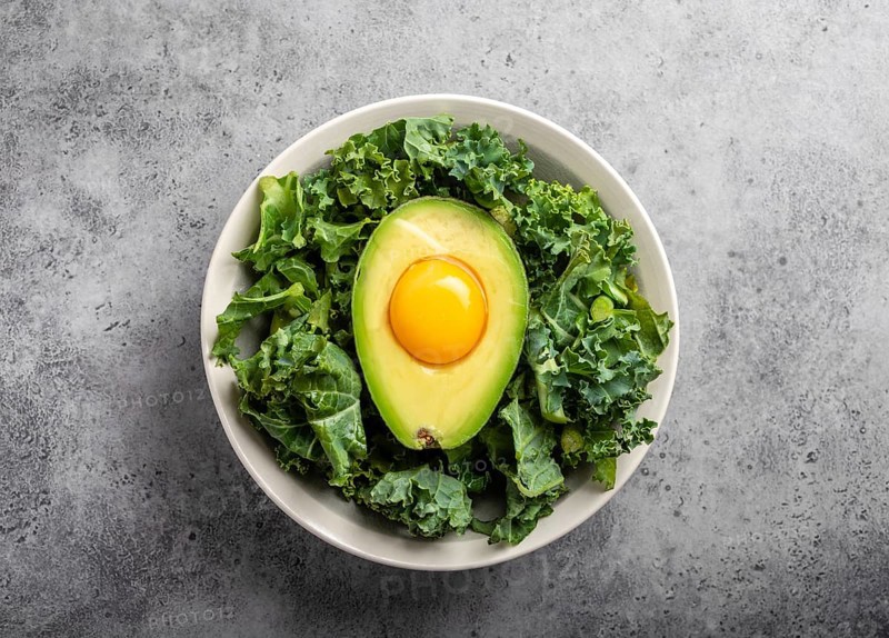 Salad bowl with kale for balanced menu, raw egg yolk in fresh cut half avocado rich in healthy fats on gray stone background. Ketogenic low carbs diet or clean eating concept
