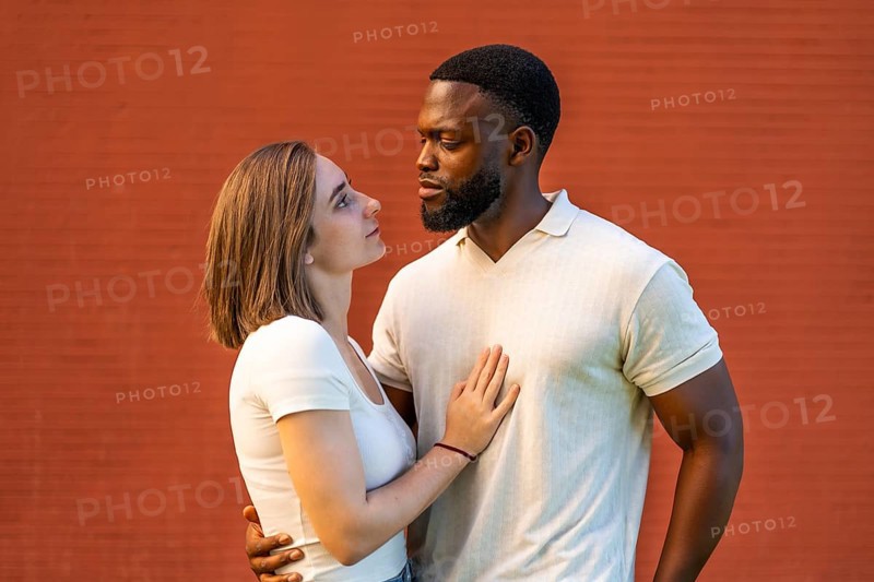 Mixed-raced couple standing embracing and looking at each other against red brick wall