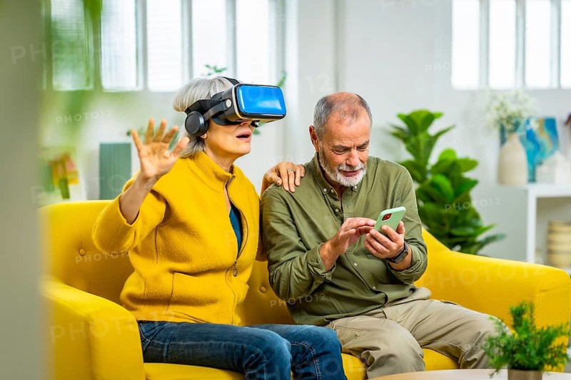 Senior woman using virtual reality glasses next to her husband sitting on the sofa using mobile app