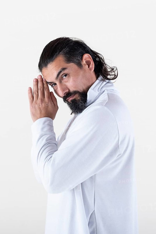 A man dressed in white clasping hands while looking at camera over white background. Tadasana yoga pose. Studio shot