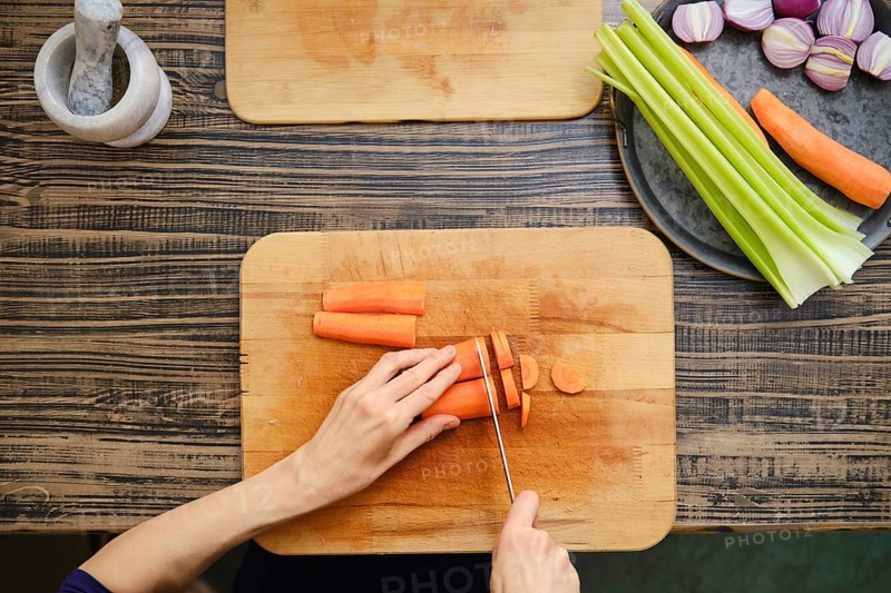 A person is chopping carrots on a wooden cutting board while surrounded by fresh vegetables, including celery and colorful onions. The atmosphere is warm and inviting, perfect for cooking