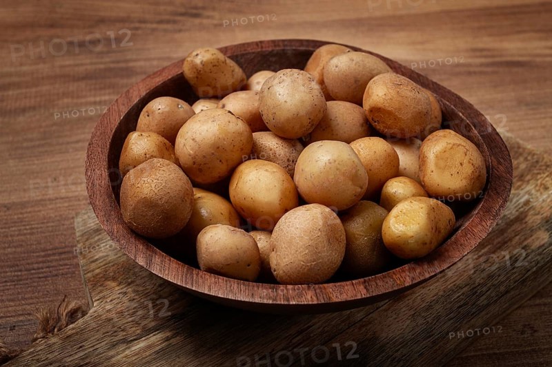 A wooden bowl filled with fresh small potatoes is centered on a rustic wooden table, showcasing their earthy tones and varied sizes in natural light