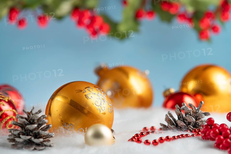 Golden and red Christmas baubles lie on artificial snow, surrounded by cones and red berries
