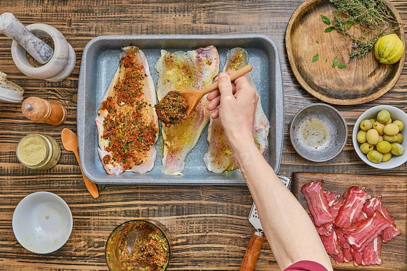 Fish fillets are laid out in a tray, being generously coated with a mix of herbs and spices. Nearby, ingredients like olives, lemon, and fresh thyme add vibrant color to the wooden table