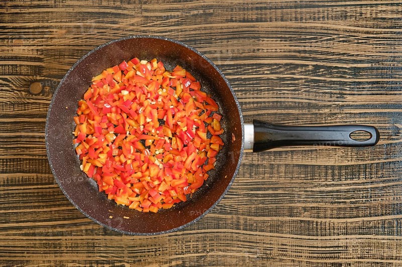 Brightly colored bell peppers are chopped and sauteing in a frying pan on a rustic wooden surface.