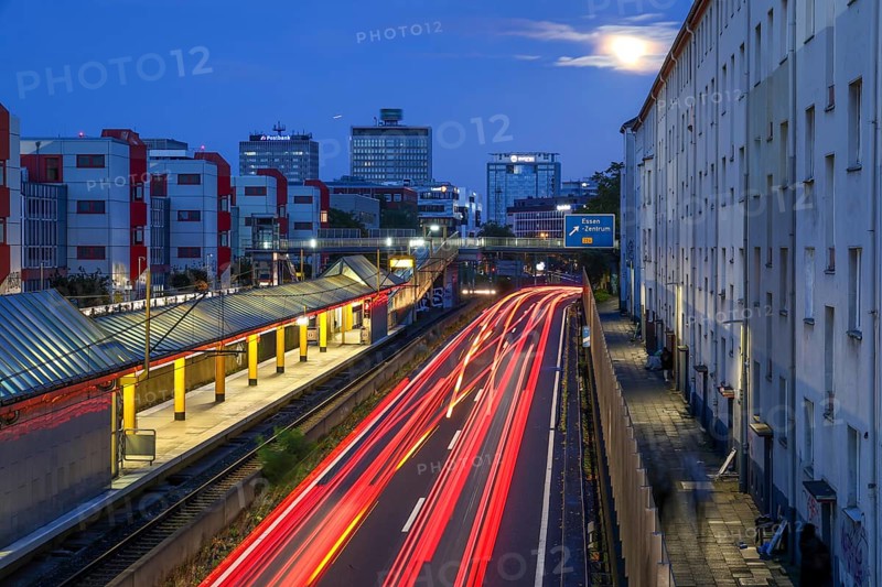 Essen, North Rhine-Westphalia, Germany, A40 motorway in the city centre at dusk. Deserted Savignystrasse underground station. On the right, residential buildings behind a noise barrier. On the wall is a LANUV air monitoring station, Essen Kruppstrasse 117 (EKRU2), on the right under the lantern. Residents are affected by noise and exhaust fumes. At the back, buildings with Postbank, Eon and Evonik lettering. Moon in the sky, Europe