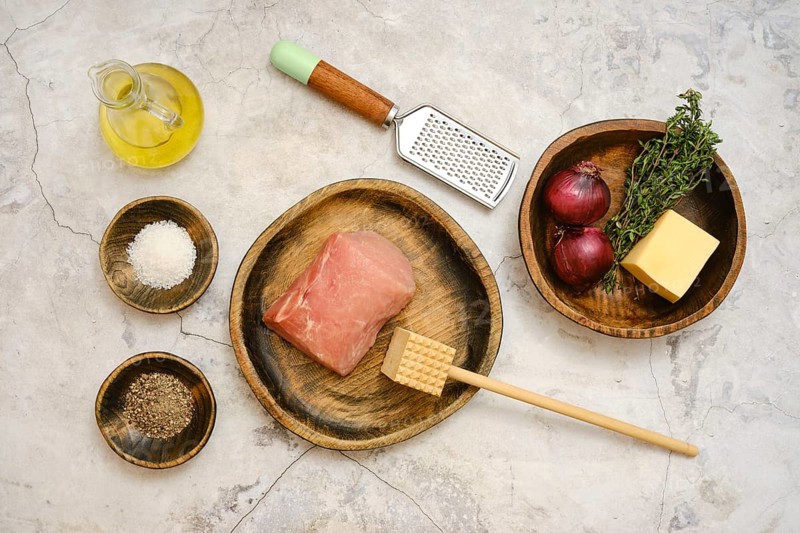 Freshly sourced pork chop is laid on a wooden plate alongside red onions, thyme, cheese, and seasoning. Cooking tools like a grater and a wooden mallet are nearby, ready for meal prep