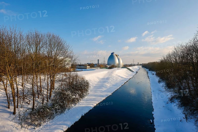 Dortmund, North Rhine-Westphalia, Germany - Sunny winter landscape in the Ruhr area, ice and snow on the renaturalised Emscher, digestion towers at the Dortmund Deusen sewage treatment plant, the treatment plant is now being equipped with a 4th purification stage, which also removes drug residues from the treated wastewater, the ecological conversion of the Emscher system consists of the construction of a central wastewater treatment system in the Ruhr area, the construction of underground sewers and the renaturation of the Emscher and its tributaries