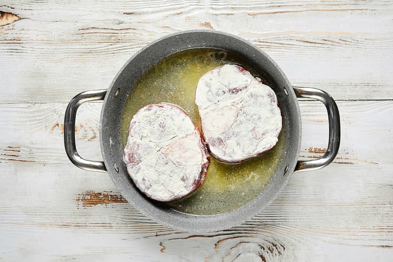 Overhead view of two floured steaks are sizzling in a pan with oil. The pan is on a white wooden tabletop