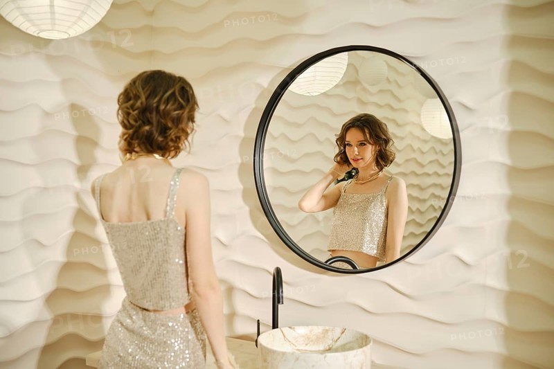 A woman dressed in a shimmering top stands in front of a round mirror, applying makeup. The stylish interior features textured walls and hanging light fixtures, setting a classy ambiance.