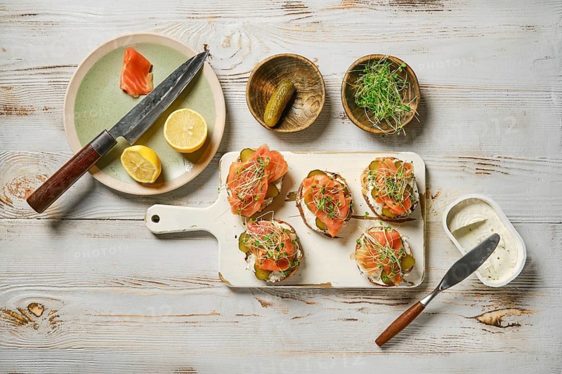 Salmon toasts topped with fresh watercress sprouts on a white cutting board. Slices of lemon and pickles complement the dish, creating an inviting rustic table setting perfect for a meal.