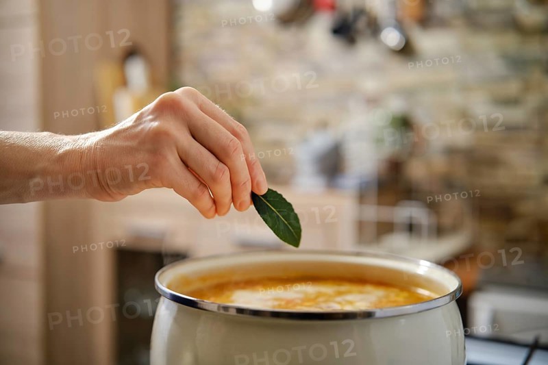 A hand reaches to place a fresh bay leaf into a pot of flavorful soup. The warm kitchen setting features wooden elements and a hint of rustic charm, creating an inviting atmosphere.