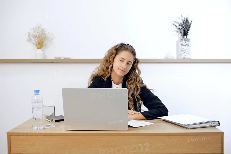 A young woman sits at a contemporary wooden desk, engaged in work on her laptop. Papers and a water bottle are placed nearby, creating a tidy workspace in a bright, minimalist office environment.