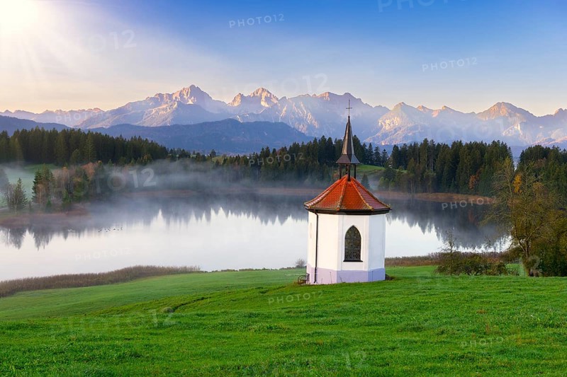Small chapel on the lake in front of an impressive mountain backdrop at sunrise, surrounded by green meadows and dense forest landscape, Hegratsrieder See near Füssen, Ostallgäu, Allgäu, Bavaria, Germany, Europe