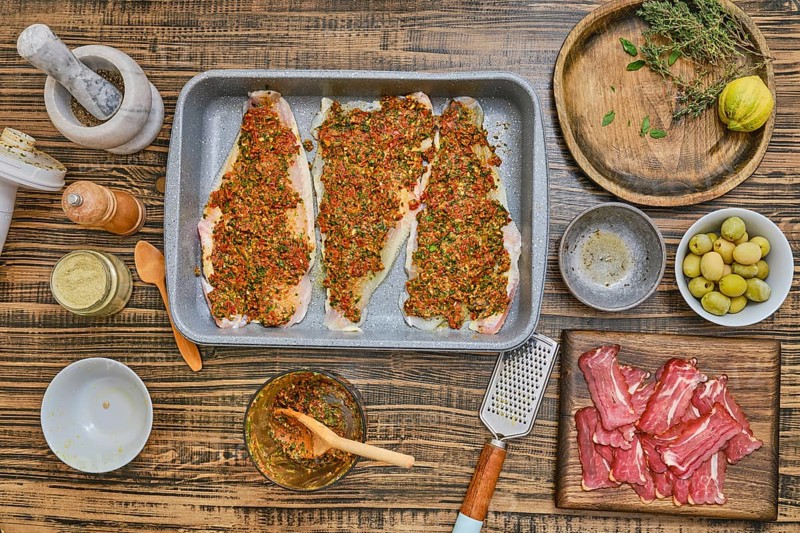 Three fish fillets are seasoned with herbs and spices, surrounded by a variety of ingredients on a rustic wooden table. Fresh olives, citrus fruit, and herbs are arranged for a flavorful meal prep